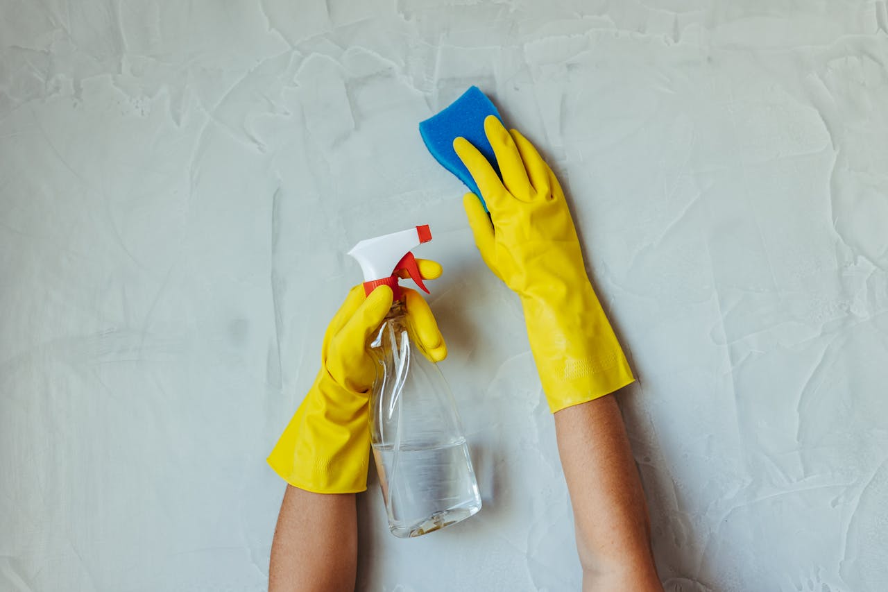 services-04 Close-up of hands in yellow gloves cleaning a wall with a spray bottle and sponge.